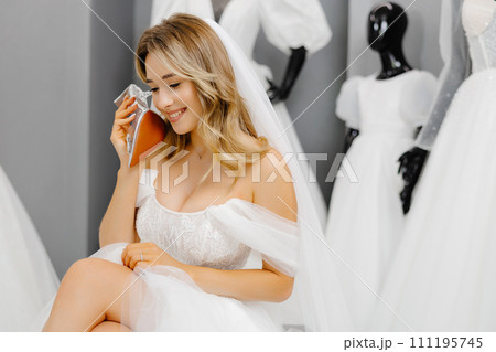 Bride-to-be wearing a wedding dress at a fitting room in a salon, choosing shoes for a wedding and having fun using a shoe as a phone Bride-to-be wearing a wedding dress at a fitting room in a salon, choosing shoes for a wedding and having fun using a shoe as a phone 111195745
