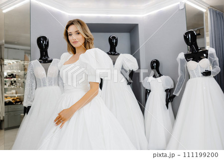 Pretty young woman is choosing a wedding dress in the shop. The bride-to-be is wearing a wedding dress for fitting 111199200