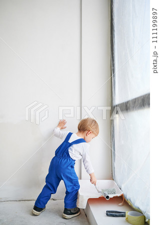 Little child construction worker standing by the window and painting white wall in apartment. Kid in work overalls using paint brush while playing at home under renovation. Little child construction worker standing by the window and painting white wall in apartment. Kid in work overalls using paint brush while playing at home under renovation. 111199897