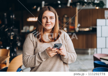 Young happy woman in glasses smiling and using smart phone in a cafe. Person using smartphone indoors. Online education, order, working or shopping concept 111202778