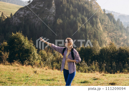 Blonde 30s girl in sunglasses pointing finger on mountains background. Young female with backpack hiking in nature. Beautiful woman walking in sport clothes. Freedom and active lifestyle concept Blonde 30s girl in sunglasses pointing finger on mountains background. Young female with backpack hiking in nature. Beautiful woman walking in sport clothes. Freedom and active lifestyle concept 111209094