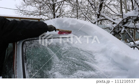 clearing snow from a car with a brush, a hand clears a thick layer of snow from the roof and windshield of a gray car, the consequences of a snow storm and snowfall for transport 111210537