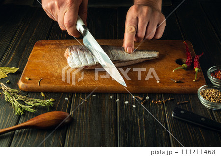 Hands of a chef using a knife to cut fresh Hake fish on a wooden cutting board. Home-cooked national fish dish according to a unique recipe 111211468