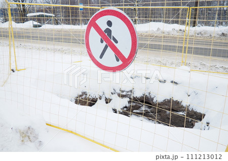 No passage sign, do not pass. Man crossed out with a red line. Round prohibitory road sign. Forbidden. A hole dug in the ground, surrounded by a metal fence. Winter, snow and snowdrifts near the road No passage sign, do not pass. Man crossed out with a red line. Round prohibitory road sign. Forbidden. A hole dug in the ground, surrounded by a metal fence. Winter, snow and snowdrifts near the road 111213012