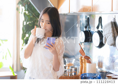 キッチンでスマホを見ながらコーヒーを飲む大学生の女性 111228413