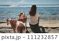 Woman with two brown dogs relax sitting on sea coast beach. Hugging dogs on the beach and looking at the ocean surf waves together. People and pets enjoying weekend time, quiet relax, beautiful nature 111232857