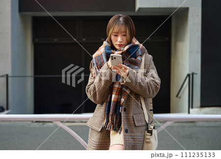スマホを持つマフラーを巻いた冬服の若い女性 111235133