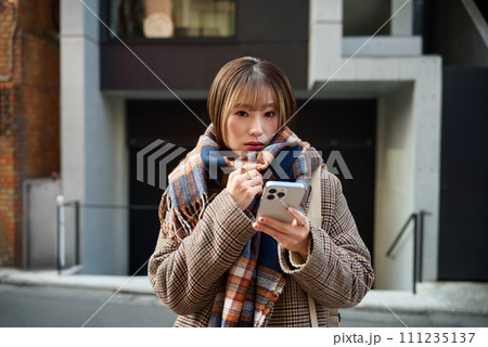 スマホを持つマフラーを巻いた冬服の若い女性 スマホを持つマフラーを巻いた冬服の若い女性 111235137