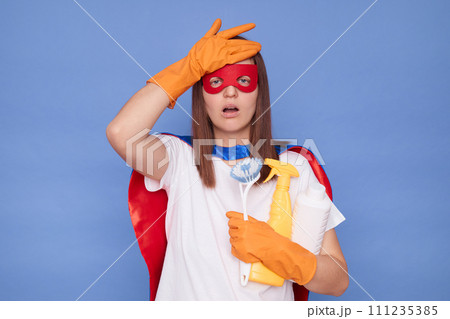 Regretful brown haired woman wearing superhero costume mask and protective rubber gloves posing isolated over blue background holding detergent and brush forgot supplies for cleanup 111235385