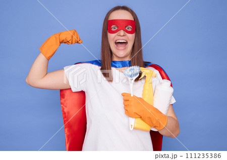 Powerful cleaning service. Joyful brown haired woman wearing superhero costume mask and orange rubber gloves embracing sanitary supplies showing her strong arm posing isolated over blue background Powerful cleaning service. Joyful brown haired woman wearing superhero costume mask and orange rubber gloves embracing sanitary supplies showing her strong arm posing isolated over blue background 111235386