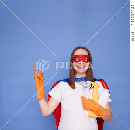 Smiling cheerful satisfied adult woman cleaner wearing gloves and superhero costume holding detergent isolated over blue background pointing away at copy space for advertisement Smiling cheerful satisfied adult woman cleaner wearing gloves and superhero costume holding detergent isolated over blue background pointing away at copy space for advertisement 111235387