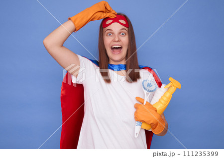 Excited adult woman cleaner wearing gloves and superhero costume raised up mask looking at camera with wow expression holding cleaning spray isolated over blue background Excited adult woman cleaner wearing gloves and superhero costume raised up mask looking at camera with wow expression holding cleaning spray isolated over blue background 111235399