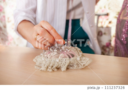 Seamstress at work. Dressmaker making clothes in modern studio. Tailor holding pencil and marking fabric. Woman standing at table with cut textile, sewing machine, thread, pins, needles, tape, cutouts 111237326