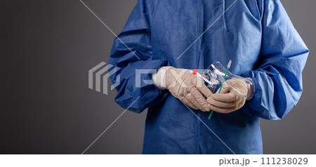 A gynecologist with a venus mirror in his hands. Gynecological accessories, brush, stick. A doctor in a medical mask, a robe, gloves, with a gynecological mirror on a gray background. A gynecologist with a venus mirror in his hands. Gynecological accessories, brush, stick. A doctor in a medical mask, a robe, gloves, with a gynecological mirror on a gray background. 111238029