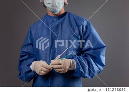 A gynecologist with a venus mirror in his hands. Gynecological accessories, brush, stick. A doctor in a medical mask, a robe, gloves, with a gynecological mirror on a gray background. 111238031