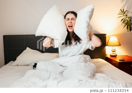 Portrait of annoyance Caucasian woman covers her ears with pillows and screams sitting in bed. Insomnia due to noisy neighbors Portrait of annoyance Caucasian woman covers her ears with pillows and screams sitting in bed. Insomnia due to noisy neighbors 111238354