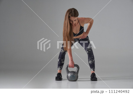 Focused sportswoman lifting heavy kettle bell, exercising in studio. Front view of strong female athlete with long hair using weight, while training, isolated on grey. Sport, weightlifting concept. 111240492