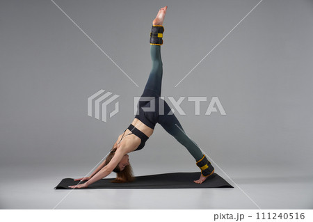 Mindful woman practicing yoga, while raising one leg in studio. Side view of graceful female with weights on legs, standing in 3-legged downward dog pose, isolated on grey. Yoga, lifestyle concept. 111240516