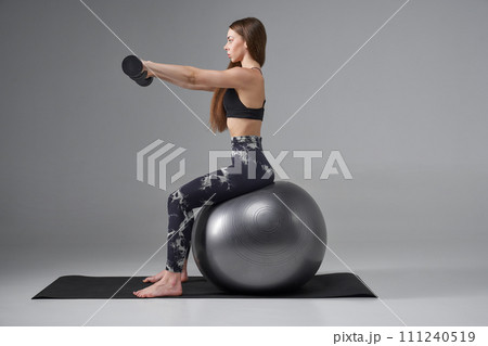 Determined woman in sports bra using dumbbells, while pumping up arms, training. Side view of female athlete working out on fitness ball with dumbbells, isolated on grey. Body care, sport concept. Determined woman in sports bra using dumbbells, while pumping up arms, training. Side view of female athlete working out on fitness ball with dumbbells, isolated on grey. Body care, sport concept. 111240519