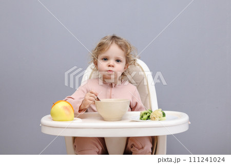 Funny little baby girl eating healthy food while sitting in high chair chewing soup looking at camera with curios facial expression isolated over gray background Funny little baby girl eating healthy food while sitting in high chair chewing soup looking at camera with curios facial expression isolated over gray background 111241024
