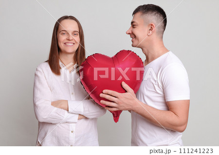 Smiling couple celebrating Valentines Day man giving present to smiling cheerful woman with heart shaped balloon expressing romantic feelings isolated over grey background Smiling couple celebrating Valentines Day man giving present to smiling cheerful woman with heart shaped balloon expressing romantic feelings isolated over grey background 111241223