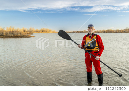 environmental portrait of a senior male paddler in a drysuit and life jacket holding stand up paddle on a shore of a lake 111242500
