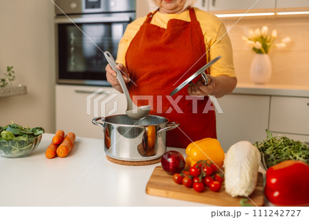 Senior 60s woman standing looking and smelling tasting fresh delicious from soup in a pot with steam at white interior kitchen Senior 60s woman standing looking and smelling tasting fresh delicious from soup in a pot with steam at white interior kitchen 111242727
