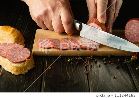 The cook cuts salami sausage on a wooden cutting board. The concept of preparing sandwiches for a snack in a restaurant kitchen 111244165