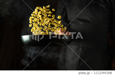 The cook cooks and throws food into a hot frying pan with noodles. Place for advertising on a black background. Pasta cooking concept or Italian cuisine 111244166