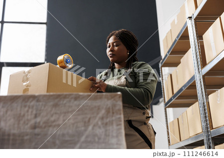 African american worker taping customers orders using adhesive tape in storehouse, preparing packages for delivery. Storage room employee working at merchandise quality control in warehouse 111246614
