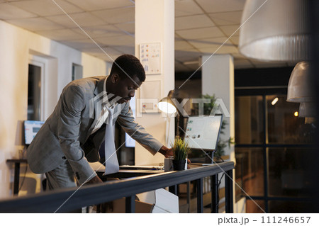 Upset employee carrying cardboard box with belongings after being dismissed from startup office. African american businessman leaving office late at night. Business management concept 111246657