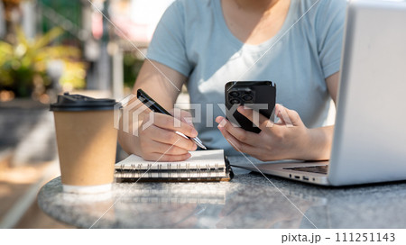A woman looking at her smartphone and taking notes in her notebook at a table outdoors. 111251143