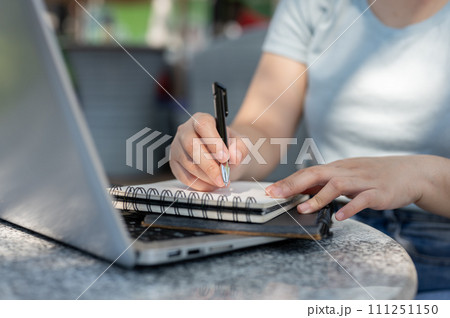 A close-up shot of a woman writing something in her notebook at an outdoor table, working remotely. A close-up shot of a woman writing something in her notebook at an outdoor table, working remotely. 111251150