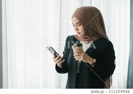 Asian Muslim businesswoman using a smartphone and enjoying coffee while standing in a modern office. 111254998