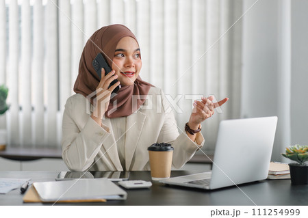 Asian muslim business woman wearing hijab using phone while working at computer in office. 111254999