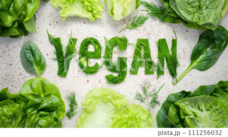 Word Vegan, title text lined up into word made from paper and green lettuce on white table with parsley, spinach broccoli, dill Word Vegan, title text lined up into word made from paper and green lettuce on white table with parsley, spinach broccoli, dill 111256032