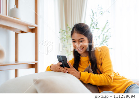 Happy young Asian woman sitting on sofa holding mobile phone using cellphone Happy young Asian woman sitting on sofa holding mobile phone using cellphone 111259094