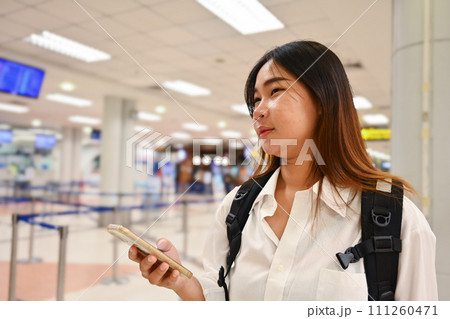 Smiling young woman checking trip destination on Internet while standing at airport Smiling young woman checking trip destination on Internet while standing at airport 111260471