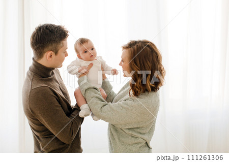 Happy couple carrying adorable baby near curtain Happy couple carrying adorable baby near curtain 111261306