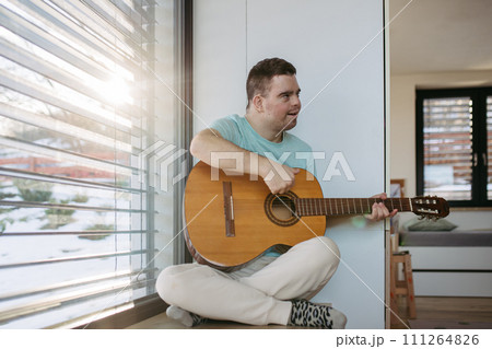 Young man with down syndrome playing acoustic guitar, sitting by window, holding and strumming guitar, making music. Young man with down syndrome playing acoustic guitar, sitting by window, holding and strumming guitar, making music. 111264826