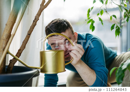 Young man with Down syndrome taking care of indoor plants, watering them, looking at camera through handle of a metal watering can. Young man with Down syndrome taking care of indoor plants, watering them, looking at camera through handle of a metal watering can. 111264843
