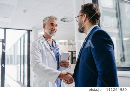 Pharmaceutical sales representative presenting new medication to doctor in medical building, shaking hands. 111264980