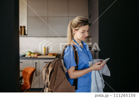 Female doctor getting ready for work, reading message on smartphone, leaving house in scrubs with backpack. Work-life balance for healthcare worker. Female doctor getting ready for work, reading message on smartphone, leaving house in scrubs with backpack. Work-life balance for healthcare worker. 111265150