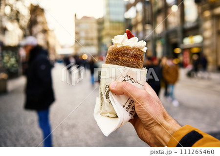 Tourist holds trdelnik against Prague street, Grilled rolled dough topped with sugar, Traditional Czech hot sweet pastry, street bakery in Prague Tourist holds trdelnik against Prague street, Grilled rolled dough topped with sugar, Traditional Czech hot sweet pastry, street bakery in Prague 111265460
