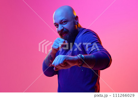 Funny fight. Gel portrait f artistic bearded bald man in blue sweater emotively posing against pink background in neon light 111266029