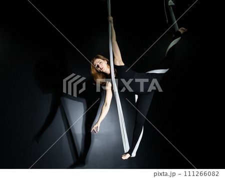 Fly yoga. Girl in a white hammock on a black background shows aerial acrobatics. Gymnastics, circus, under dome. Play of light and shadow creates feeling of a theatrical performance. Sports activity 111266082