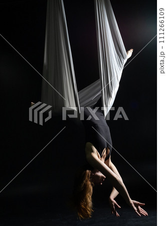 Fly yoga. Girl in a white hammock on a black background shows aerial acrobatics. Gymnastics, circus, under dome. Play of light and shadow creates feeling of a theatrical performance. Sports activity Fly yoga. Girl in a white hammock on a black background shows aerial acrobatics. Gymnastics, circus, under dome. Play of light and shadow creates feeling of a theatrical performance. Sports activity 111266109