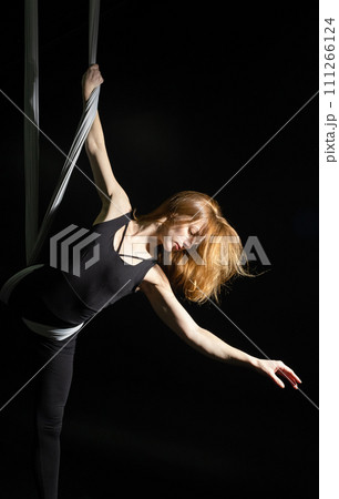 Fly yoga. Girl in a white hammock on a black background shows aerial acrobatics. Gymnastics, circus, under dome. Play of light and shadow creates feeling of a theatrical performance. Sports activity 111266124