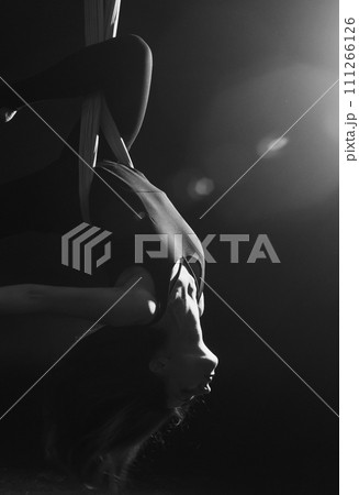 Fly yoga. Girl in a white hammock on a black background shows aerial acrobatics. Gymnastics, circus, under dome. Play of light and shadow creates feeling of a theatrical performance. Sports activity Fly yoga. Girl in a white hammock on a black background shows aerial acrobatics. Gymnastics, circus, under dome. Play of light and shadow creates feeling of a theatrical performance. Sports activity 111266126