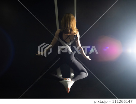Fly yoga. Girl in a white hammock on a black background shows aerial acrobatics. Gymnastics, circus, under dome. Play of light and shadow creates feeling of a theatrical performance. Sports activity 111266147